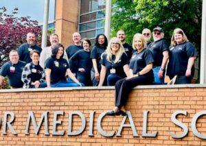 Group of UAMS staff members posing together outdoors on a brick sign in front of a campus building, wearing coordinated dark shirts and smiling, representing teamwork and employee engagement.
