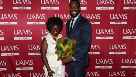 A man and woman stand in front of a UAMS red banner. The man is holding an award.