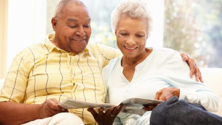 An older couple sits on a couch reading a brochure together.